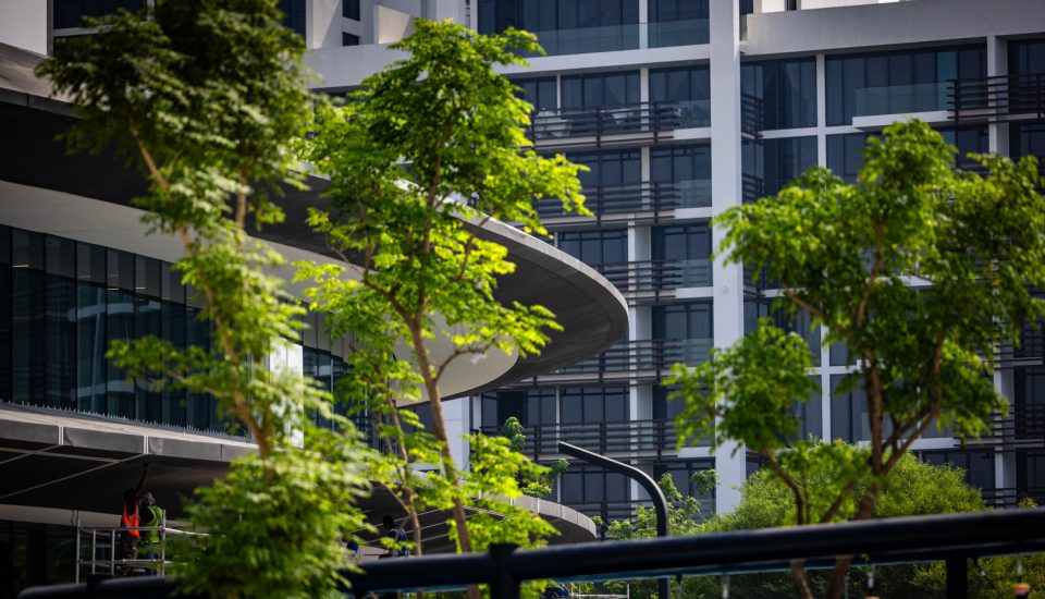 The image showcases a modern urban scene dominated by contemporary architecture. In the forefront, lush green trees with their branches and leaves partially obscure the view, lending a refreshing touch of nature to the otherwise man-made environment. The main architectural structure in the frame appears to be a multi-storied building, exhibiting a mix of dark-tinted glass windows and concrete panels. Unique design elements include a circular, overhanging balcony or platform, which extends from one of the building sections. The building's facade features a combination of vertical and horizontal lines, possibly balconies or exterior design elements. In the lower part of the photo, there are several workers dressed in safety vests, operating on some form of elevated platform, possibly engaged in maintenance or construction activities. The overall ambiance suggests a dynamic urban setting, where nature seamlessly intermingles with modern design.